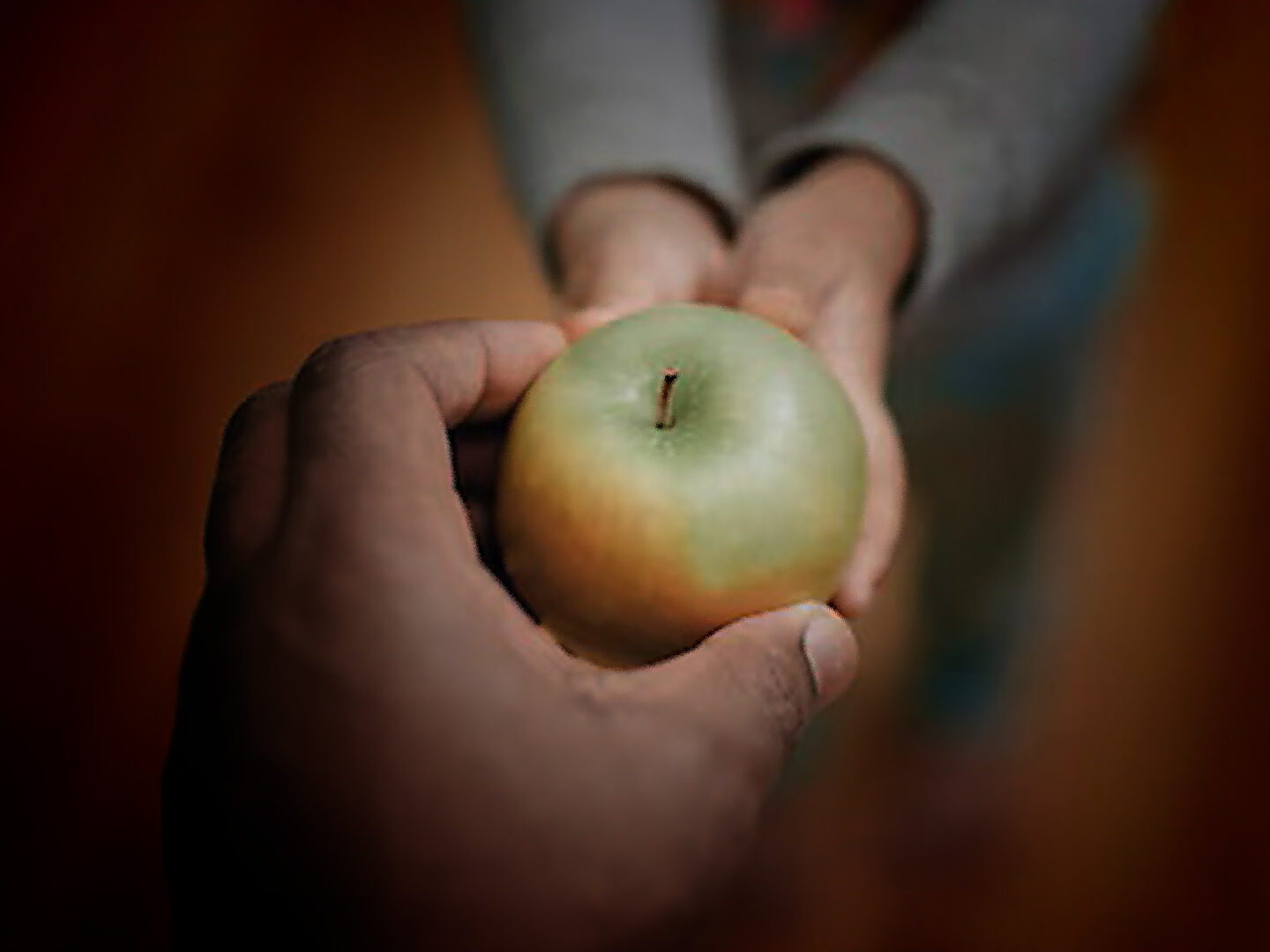 A hand holding a green apple is being offered to a child's hand in a warm, indoor setting.